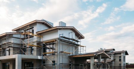 Casa de dois pavimentos em construção, cercada por andaimes metálicos, com estrutura e reboco em andamento.