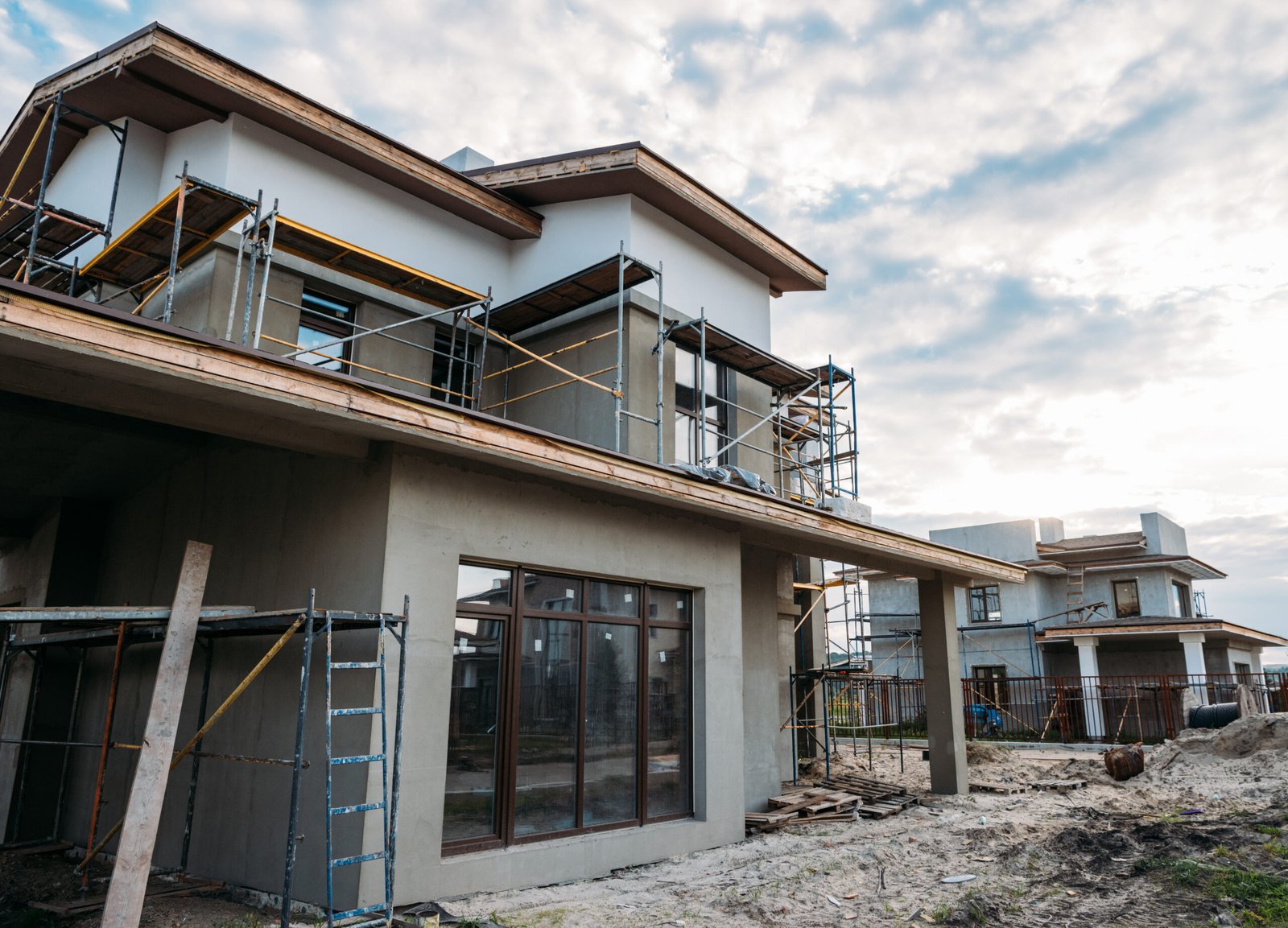 Casas modernas em construção com andaimes e janelas instaladas, etapa de reboco em andamento.